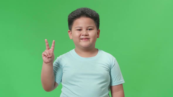 A Smiling Asian Little Boy Showing Gesture Peace While Standing On Green Screen In The Studio