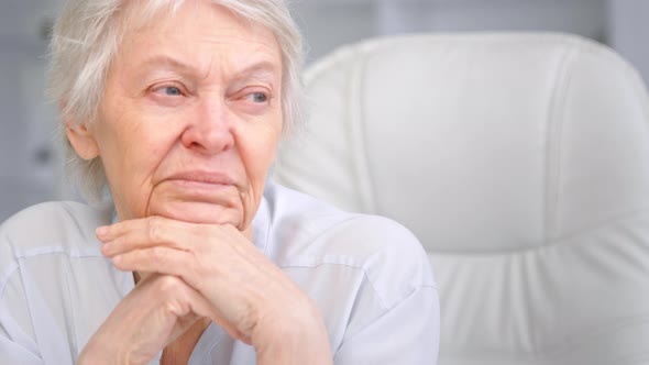 Thoughtful grey haired senior lady looks into window holding head over hands