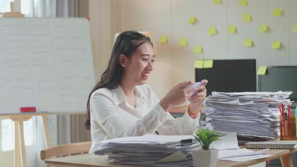 Asian Woman Enjoys Playing Game On Smartphone After Working With Documents At The Office