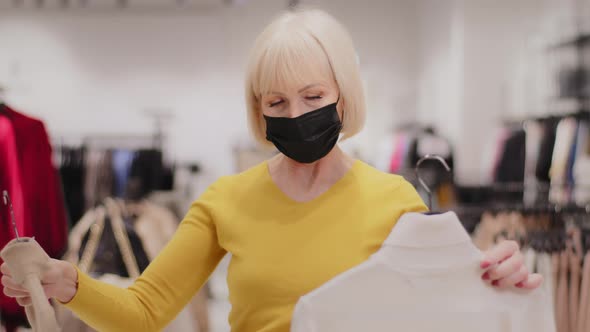 Mature Elegant Woman Shopper in Medical Mask Stands in Clothing Store Chooses Clothes Holds Two