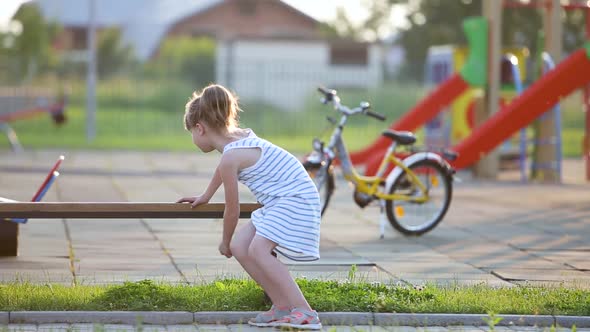 Cute Little Girl Sitting on a Bench and Riding a Bicycle on a School Yard in Summer