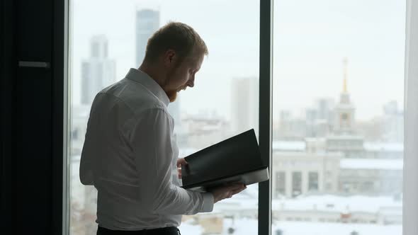 a man with documents stands by the window overlooking the city