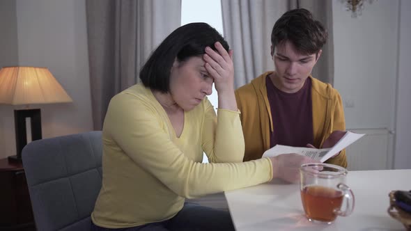 Portrait of Strict Dissatisfied Caucasian Mother Sitting with Teenage Son and Checking List with His