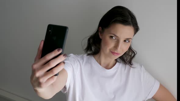 Woman in a White T-shirt on a Light Wall Makes a Selfie