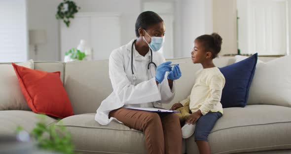 African american female doctor wearing face mask measuring temperature of girl at home
