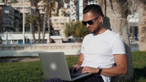 Young Bearded Man Tshirt Wearing Glasses Sitting on the Grass in the Park with His Laptop and