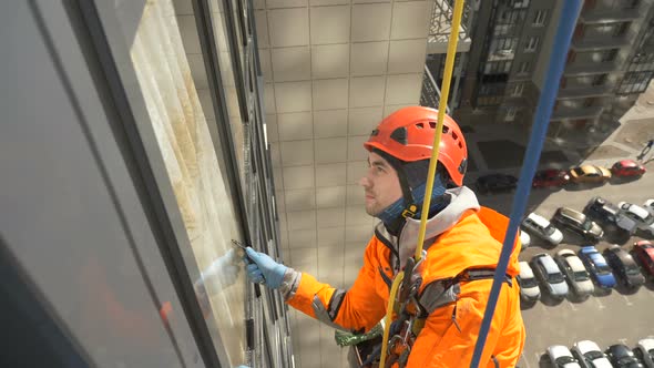 Professionally Equipped Industrial Climber in Helmet and Gloves Washes Windows with a Screed