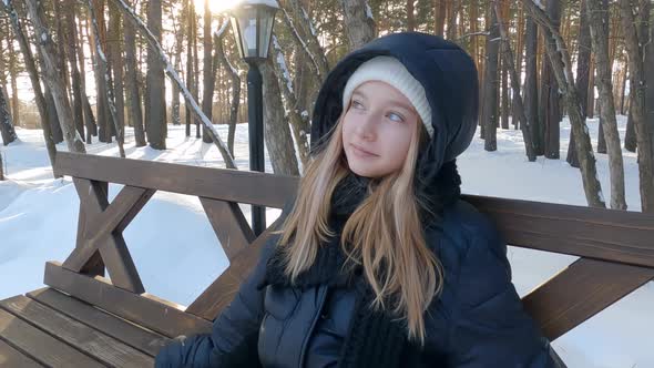 Thoughtful Pretty Blonde Girl in Black Coat Sitting on Wooden Bench in Winter Snowy Park Enjoying