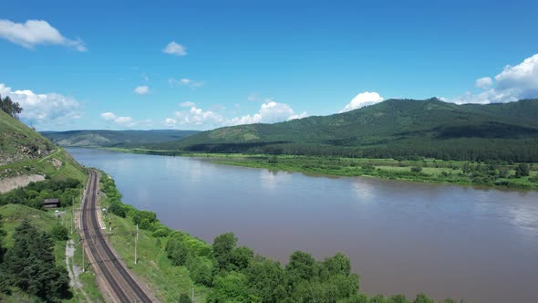 Flying Above a Glacial River in Siberia Russia with Green Mountains and Railway Track in the