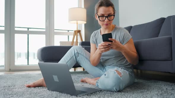 Casually Dressed Woman Sitting on Carpet with Laptop and Smartphone and Working in Cozy Room