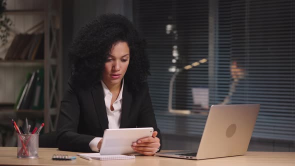 Portrait of African American Woman Views Information on Digital Tablet and Laptop Verifies Data