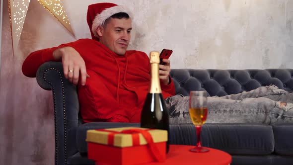 Young Man in Santa Claus Hat Browses Smartphone and Drinks Champagne Lying on Couch