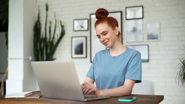 Smiling Young Redhead Woman Is Typing on Laptop at the Desk at the Home Office.