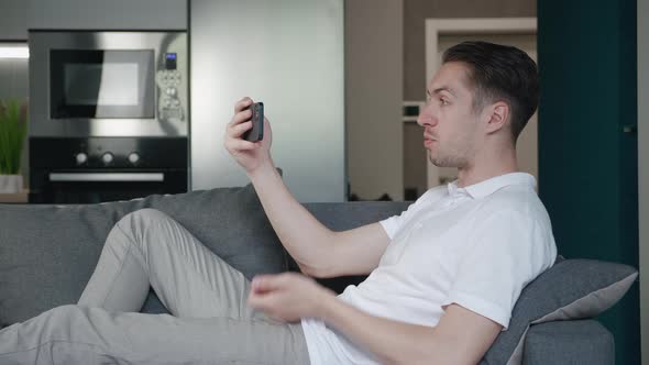 Smiling Young Man with Smartphone Making Video Call While Lying on Couch in Living Room