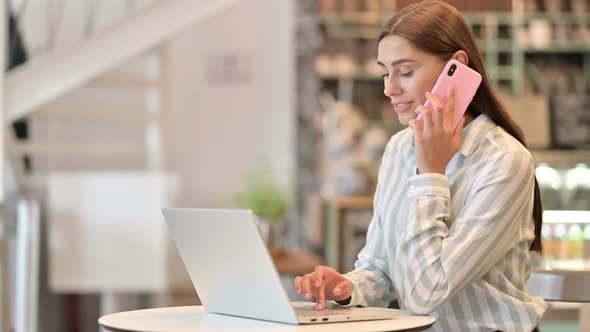 Young Latin Woman with Laptop Talking on Smartphone in Cafe