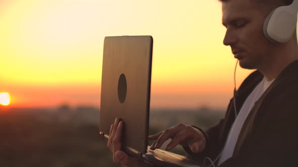 A Man Freelancer in Headphones Standing on the Roof at Sunset Writes on the Keyboard Code Pages