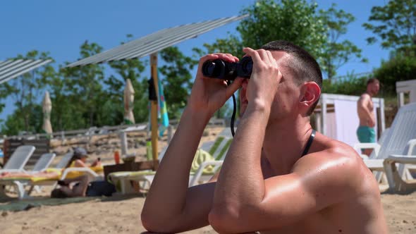 Man on the Beach in Swimming Trunks is Watching Someone Through Binoculars