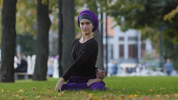 Young Girl Muslim Woman in Hijab Sits on Green Grass in Park Takes Leg By Foot Bends Turns to Side