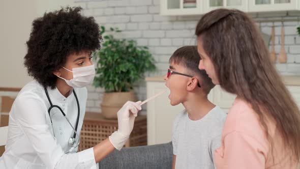 Afro American Woman Doctor in Mask Looking at Child's Throat While Sitting on Sofa at Home