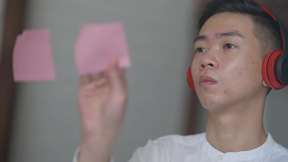 Headshot Portrait of Serious Concentrated Chinese Man in Headphones Sticking Sticky Notes on Mirror