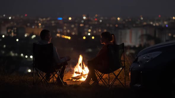 Young couple in love talking with each other sitting on chairs near bonfire at night enjoying