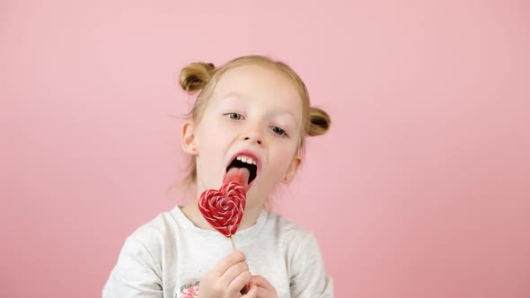 Funny Little Blonde Girl Smiling and Licking Red Heart Shape Lollipop Caramel on Pink Background