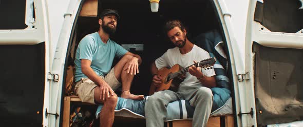 Two men sitting in back of a van singing and playing the guitar