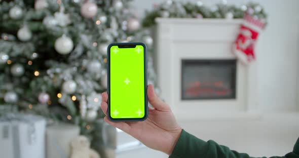 Man Hand Holding the Black Smartphone with Green Screen on Christmas Decorated Background. Close-up