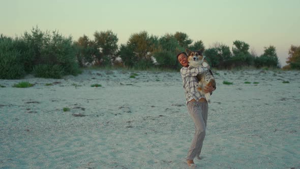 Happy Laughing Woman Holding Cute Welsh Corgi Dog on Hands and Walking on Sandy Beach at Sunset