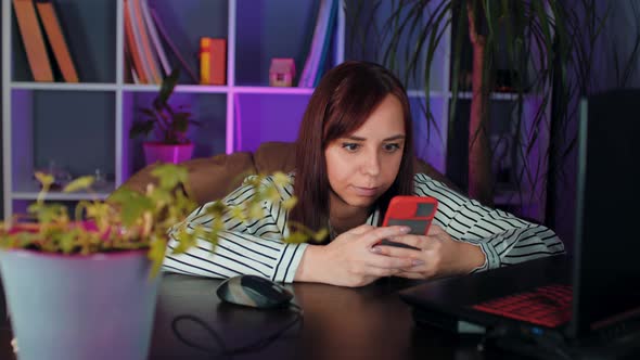 Young Woman Using Smartphone Sitting in Armchair Before Computer in Office