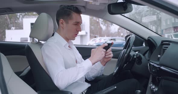 Business Man in White Shirt Excitedly Plays a Game on His Smartphone While Sitting in the Drivers