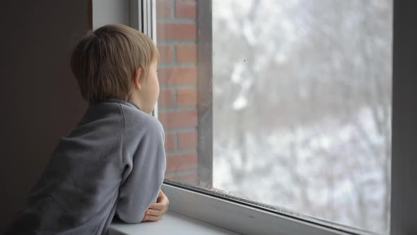 A Little Boy Looks Through the Window on a Heavy Snowfall Waiting for Christmas To Come. Slowmotion