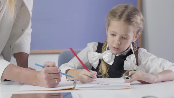 Portrait of Charming Caucasian Schoolgirl Writing with Red Pencil While Unrecognizable Teacher