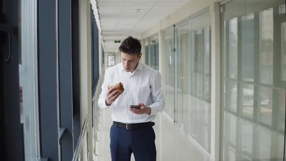 Young Man Types on the Mobile Phone and Eats Burger in Corridor of Office Centre