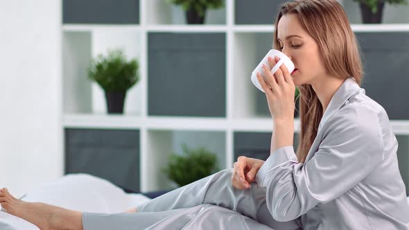 Pleasant Young Morning Woman Drinking Hot Beverage From Mug Sitting on Bed