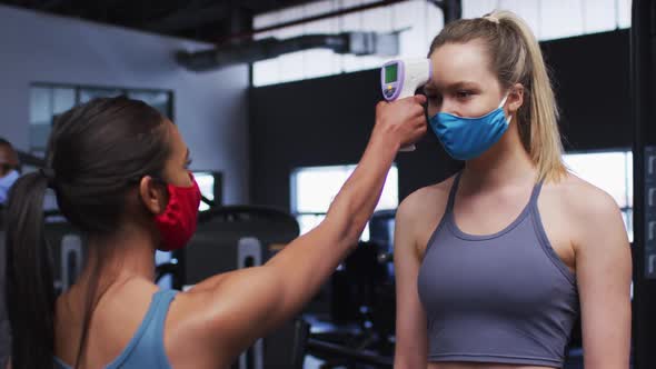 Fit caucasian woman wearing face mask measuring temperature of fit caucasian woman in the gym