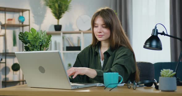 Young Girl in Casual Clothes Sitting at Her Workplace in Home Office and Working on Computer