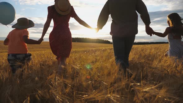 Happy Family with Two Children Holding Hands of Each Other and Running Through Wheat Field at Sunset