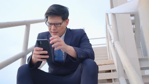 Business man with suit sit on step or staircase on rooftop of high building and use mobile phone