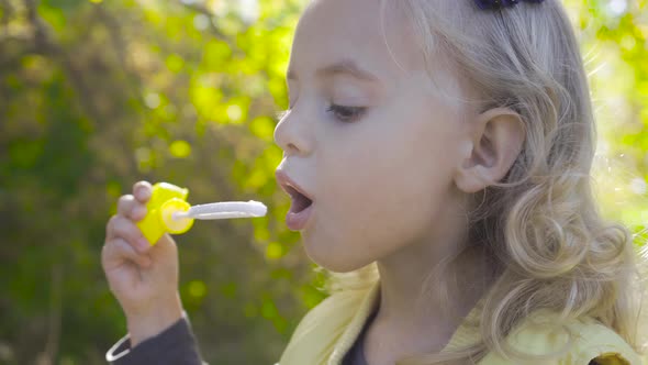 Side View of a Cute Blond Girl Blowing Soap Bubbles and Smiling. Little Charming Child with Brown
