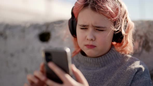 Cute Teen Sad Sitting Outside on a Sunny Day and Listening to Music in Wireless Headphones Via the