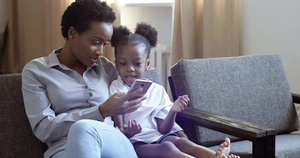 Mixed Race Mom with African Black Cute Daughter Sitting on Couch with Mobile Device Smartphone