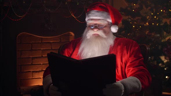 Bearded Senior Man in Santa Clause Outfit Flipping Through Pages of Book with Red Cover in