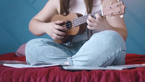 Young Woman Composer is Sitting on Bed with Ukulele in Her Hands and Writing Down Her Thoughts and