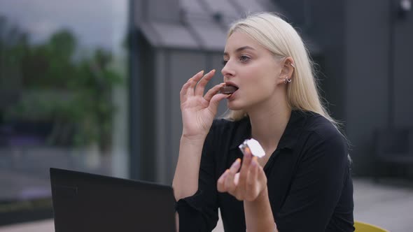 Young Slim Blond Woman Eating Sweet Delicious Candy Sitting on Office Terrace Outdoors