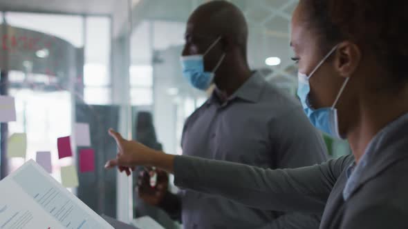 Diverse male and female business colleagues in face masks discussing by transparent board in office