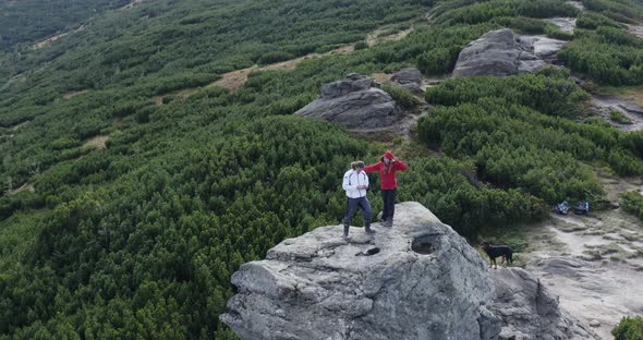 a guy and a girl are standing on a rock in the mountains with a big wind next to a black Rottfeiler