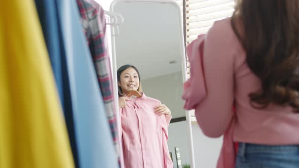 Asian lady choosing clothes on clothes rack dressing looking herself in mirror in bedroom.