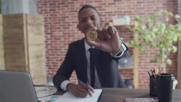 Black Man in a Strict Suit Smiling Shows a Bitcoin Coin with Hand and Begins to Write Pen on Paper