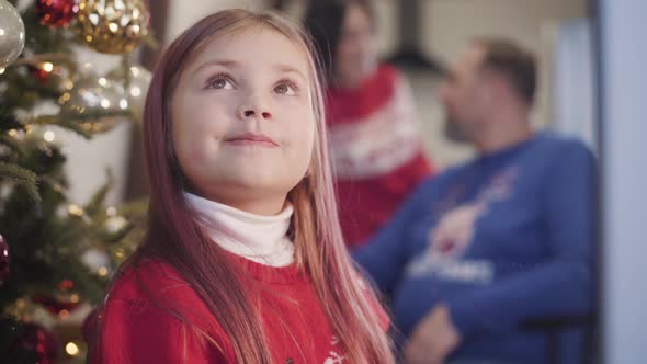 Portrait of Charming Caucasian Girl Looking Out the Window on Christmas Eve As Blurred Parents